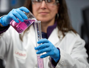 Female laboratory technician does tests, wearing uniform and protection glasses. She is holding vials with pink colored liquids