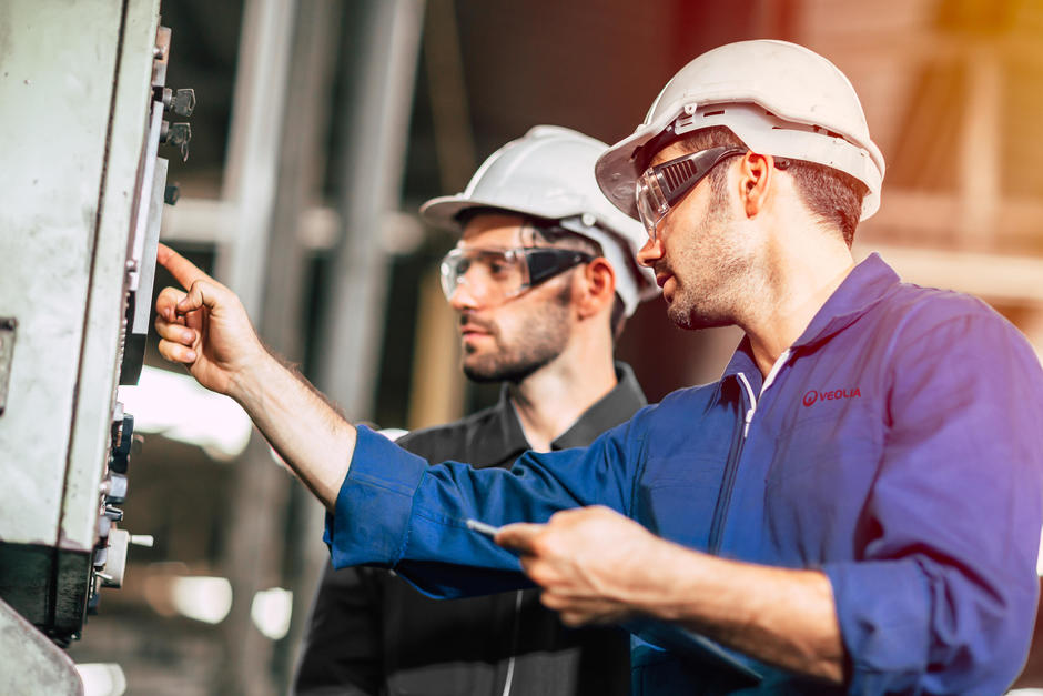 Two man wearing safety helmets, glasses and jumpsuits working in a factory.